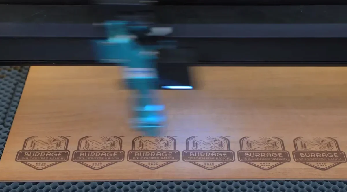 Laser engraving machine etching 'Burrage Wilderness Ranch' logos on a wooden surface.