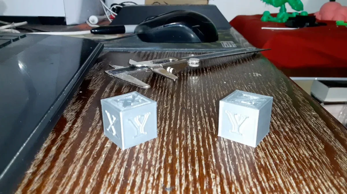 Two gray 3D printed calibration cubes with 'X' and 'Y' on a wooden desk near calipers.