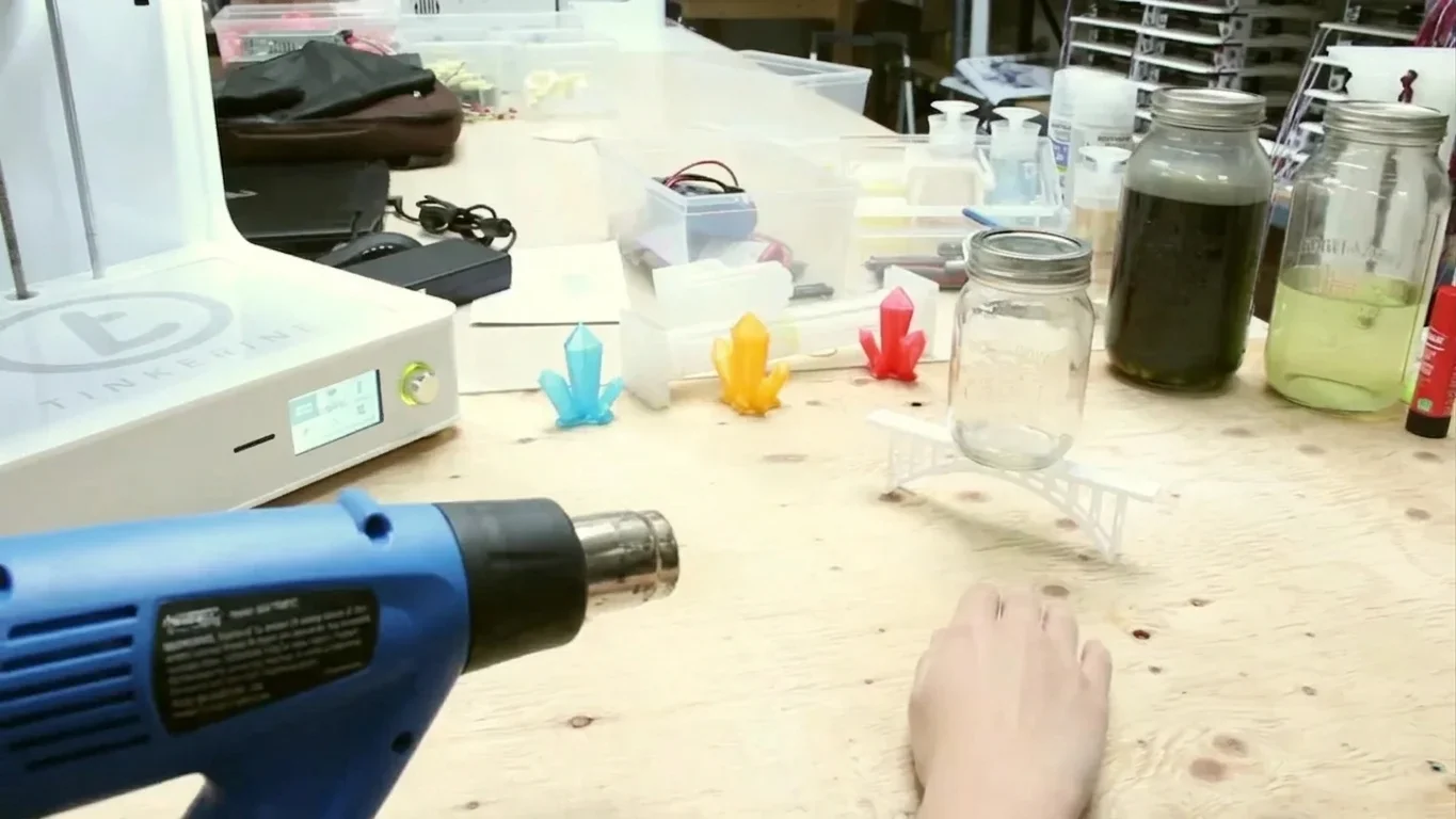 3D print setup with heat gun demonstrating PLA warping prevention techniques.